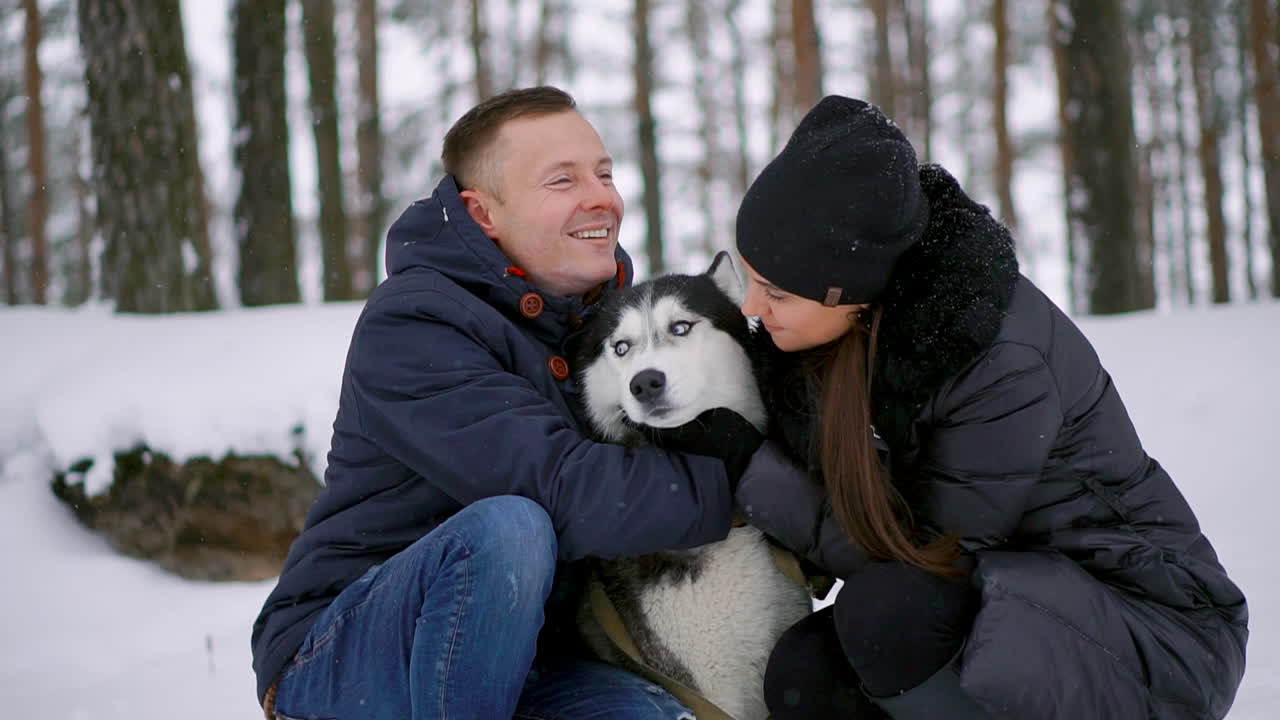 Family portrait of cute happy couple hugging with their alaskan malamute dog licking man's face. Funny puppy wearing santa christmas deer antlers and kissing woman. Freedom lifestyle pet lovers.