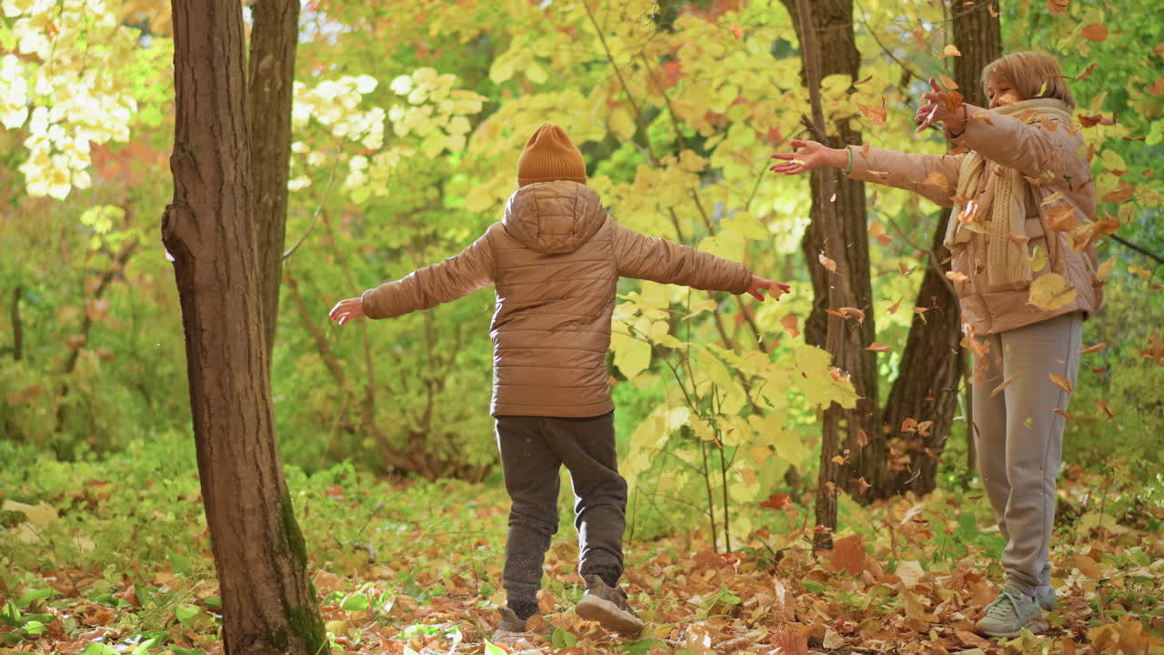 woman joyfully tossing autumn leaves into air while daughter spins around in golden forest, both smiling brightly and sharing playful moment surrounded by rich fall foliage
