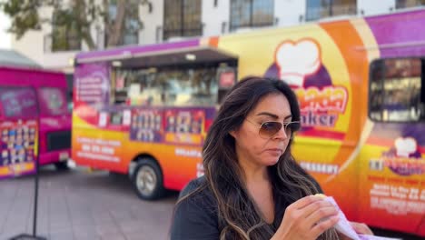 attractive hispanic woman eating a snack purchased from a food truck in the blurred background