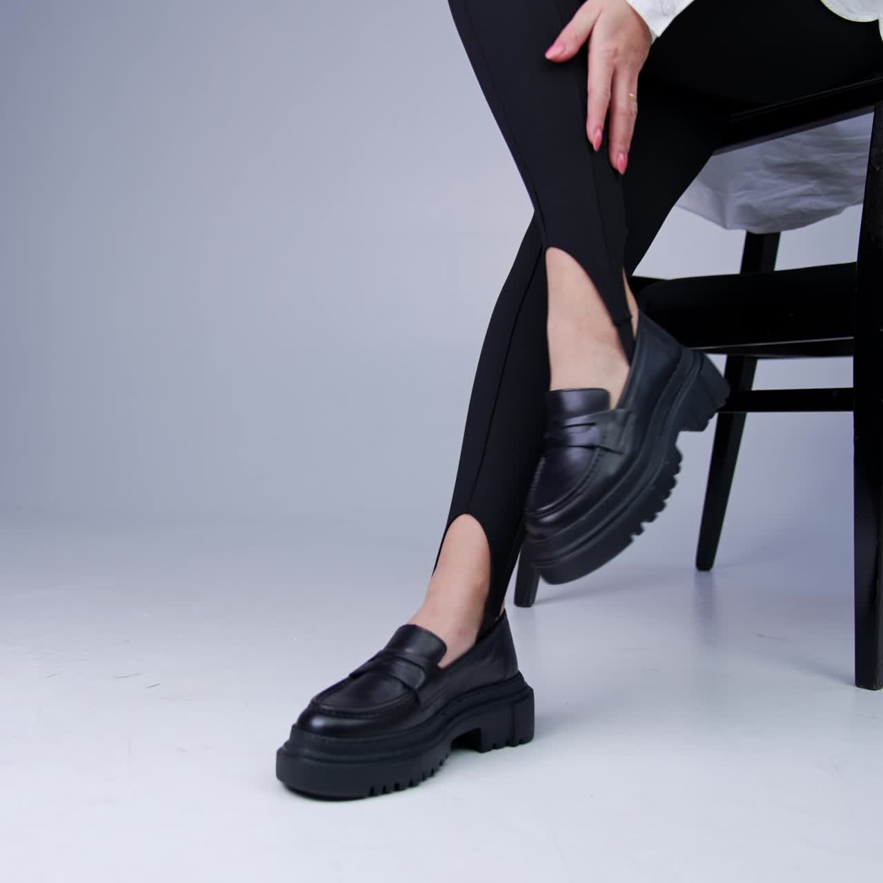Unrecognized woman wearing black leggings and white shirt demonstrates her modern footwear. Model sits on the chair touching her black stylish tractor soles shoes