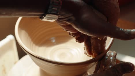 a close-up of a pottery teacher's hands guiding a student's hands to shape a wet clay bowl on the potter's wheel, teaching the craft in a creative workshop.