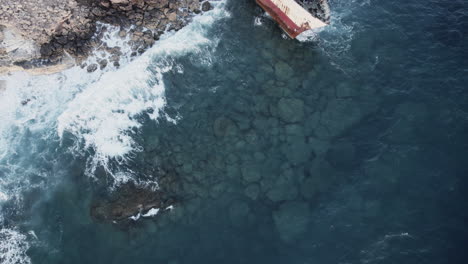 A-dramatic-aerial-top-view-of-the-Edro-III-shipwreck-lying-on-the-rocky-coast-of-Cyprus,-highlighting-the-contrast-between-the-rusted-ship-and-the-powerful-waves