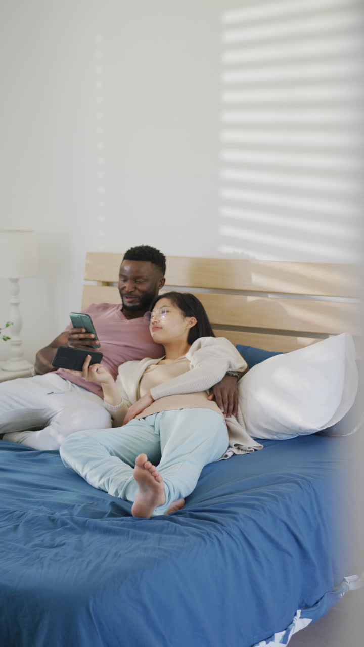 Vertical video of happy diverse couple lying and using smartphone in bedroom