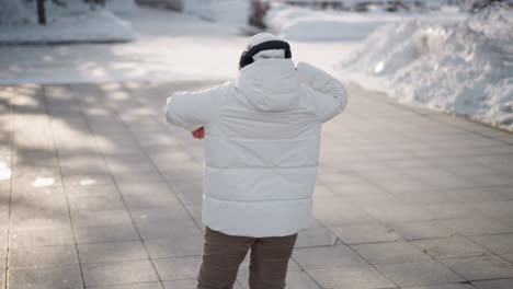 back view of dancer wearing white puffer coat, beanie and headphones practicing fluid dance moves on tiled plaza lined with snowbanks and bare trees under clear cold winter daylight in urban park
