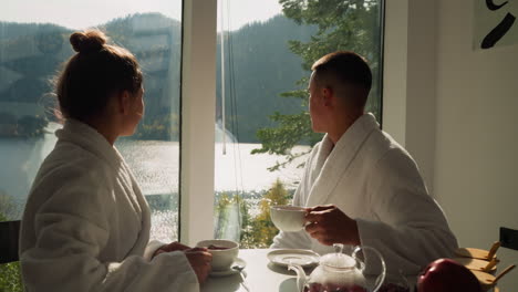 newlyweds admire nature looking in window. couple in terry bathrobes enjoys aromatic tea in glamping hotel. concept of luxury weekend in wild nature area