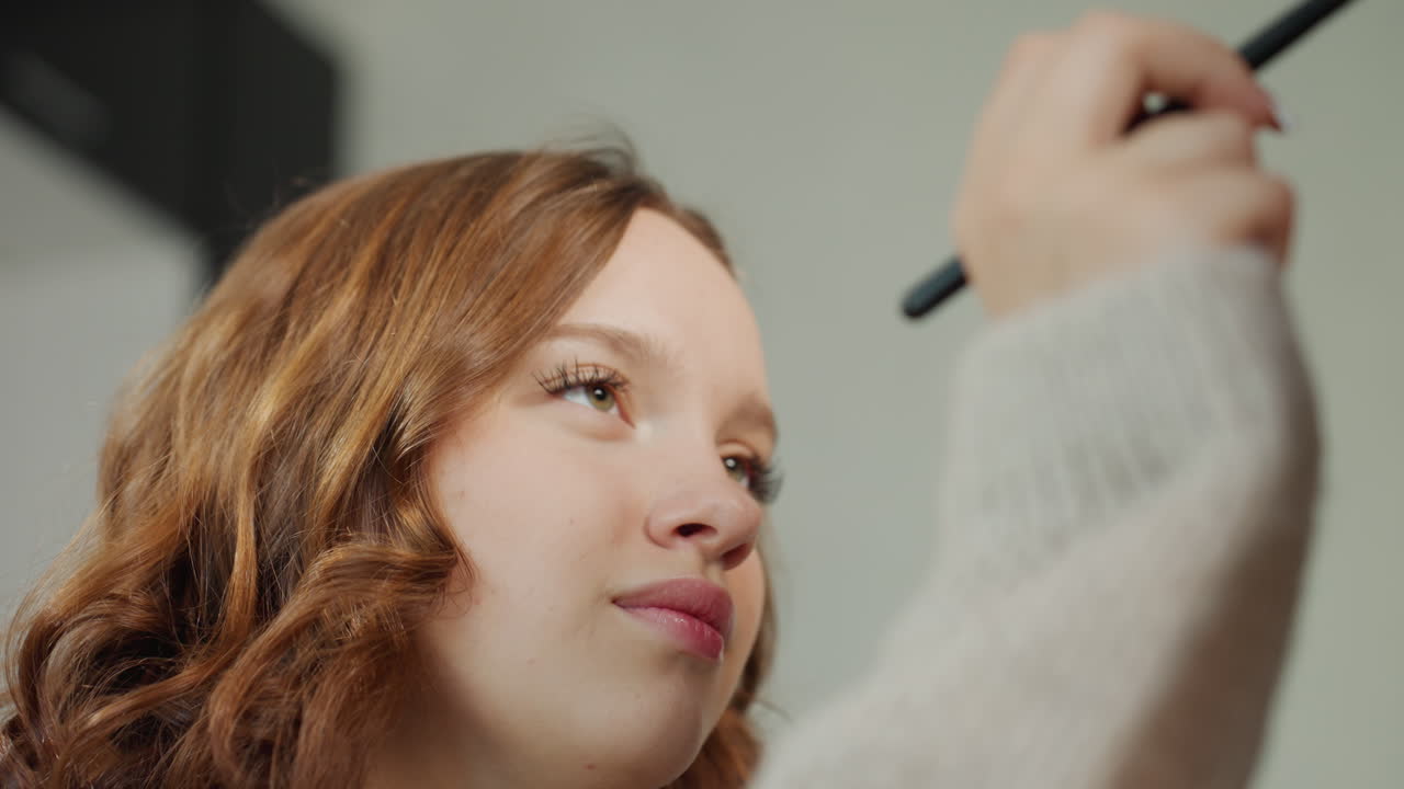 CloseUp Girl Receiving Eye Makeup Application In Cozy Studio Setting, Redhead Sits As Makeup Artist Gently Applies Shadow With Brush, Soft Lighting And Focused Expression, Sweater Sleeve Visible