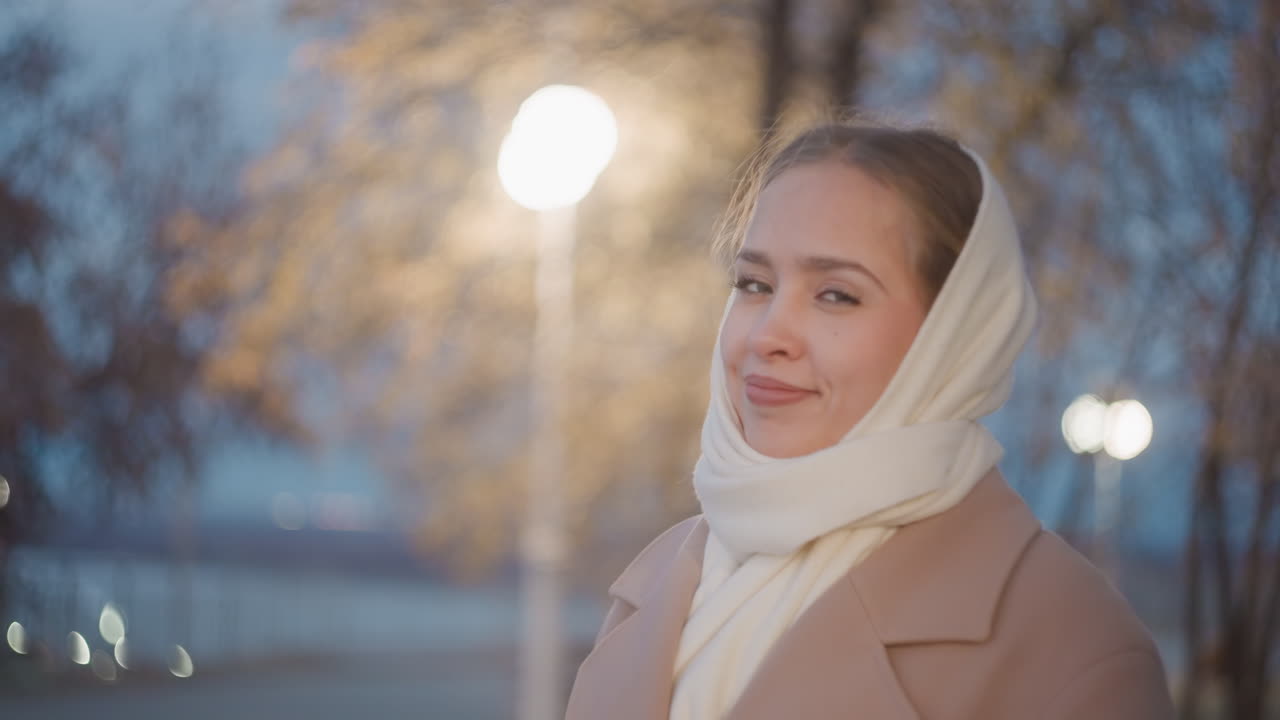 Vrouw met sjaal loopt door de duisternis van het park. Jonge dame staat rustig onder gloeiende straatlantaarns in het park. Jonge vrouw met sjaal pauzeert vredig onder straatlantaarns in het park tijdens de schemering.