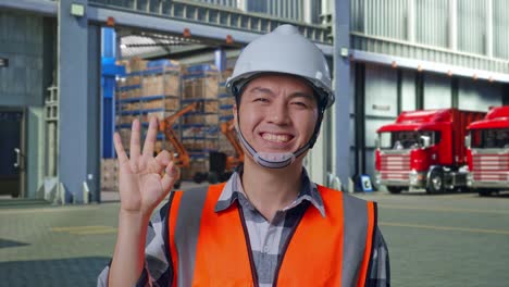 close up of asian male engineer with safety helmet smiling and showing okay gesture to the camera while standing , outside of logistics distributions warehouse