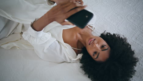 gentle lady tapping smartphone bedroom. african american curly girl laying bed