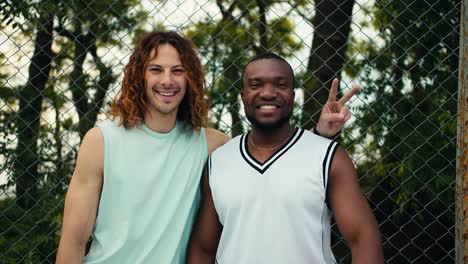 retrato de dos amigos de cabello rojo, hombre de cabello rizado y un hombre negro en una camiseta blanca posando y mirando a la cámara y sonriendo contra el fondo de una valla de malla en una cancha de bucksball
