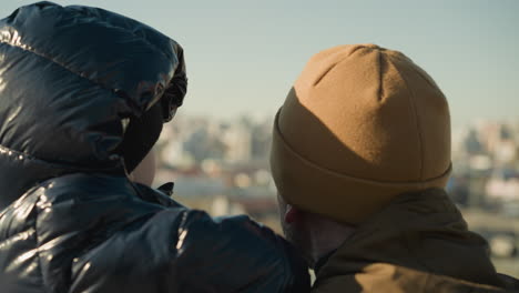 a father carrying his son looks out over a cityscape, while the son glances looks at the camera before turning away, both are dressed in jackets and beanies, with the blur cityscape