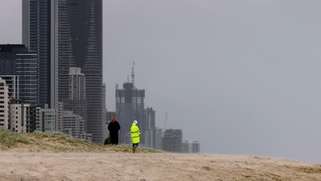 a view of skyscrapers near a sandy beach with two people in high-visibility jackets.