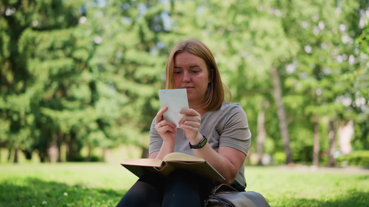Student sitting in sunny park turning pages of book discovering hidden picture, gazing at it curiously under green trees, surrounded by peaceful summer atmosphere filled with focus