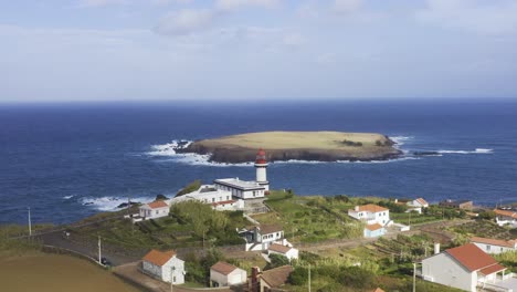 pueblo costero rural con un faro y una isla en el océano atlántico, cielo soleado y nublado en topo, isla de são jorge, azores, portugal