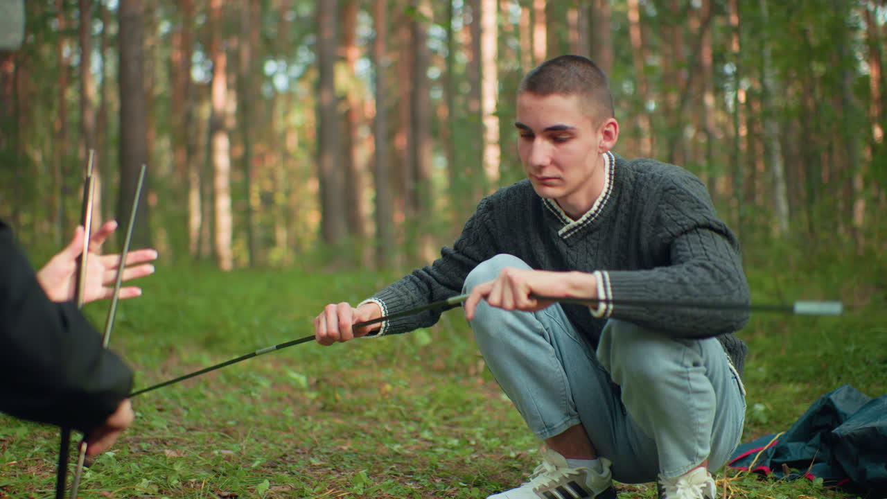 Lady hands tent pole to husband who squats to fix it during forest camping while she playfully holds next pole back teasingly, both smiling and enjoying nature surrounded by tall trees and equipment