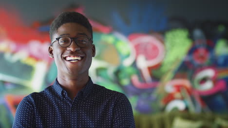 young black male creative smiling to camera in front of mural in casual office, head and shoulders