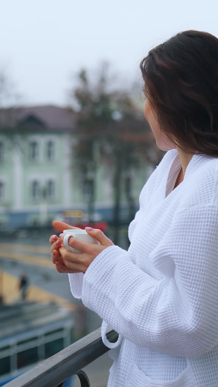Photo of a beautiful girl in a bathrobe on the hotel balcony with a cup of coffee. The morning of the bride. A young woman is having breakfast on the balcony. Vertical video