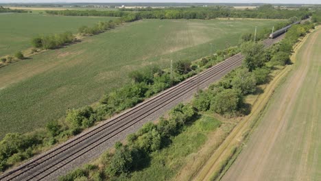 an aerial view shows double railway tracks cutting through lush green farmland bordered by trees and vegetation. the drone record a moving train.
