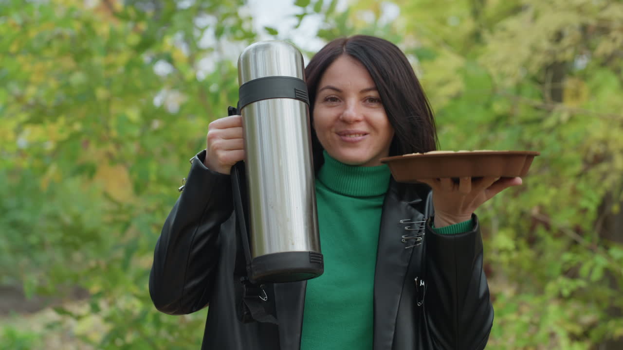 Woman holding thermos and pie outdoors, smiling at camera among green foliage, wearing green sweater and leather jacket, offering stainless steel flask and wooden plate, autumn park setting, casual