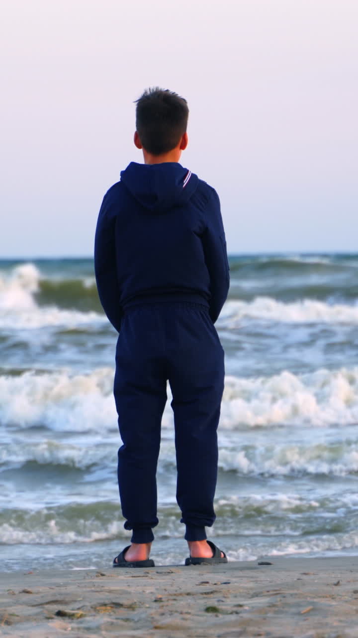 Teenage boy looking at the sea. Backside view of a child in clothes standing on a sandy beach near the sea and looking on waves in summer evening. Vertical video