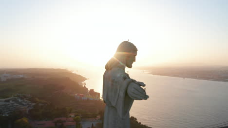 Close-up-aerial-orbit-of-Sanctuary-of-Christ-the-King-statue-on-the-hill-with-reveal-of-Ponte-25-de-Abril-red-bridge-in-Lisbon,-Portugal