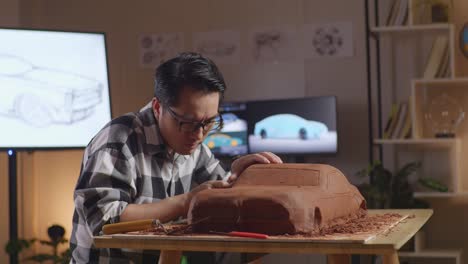 asian man automotive designer works on the sculpture of car clay using rake or wire to smooth out the surface and create details in the design in the studio