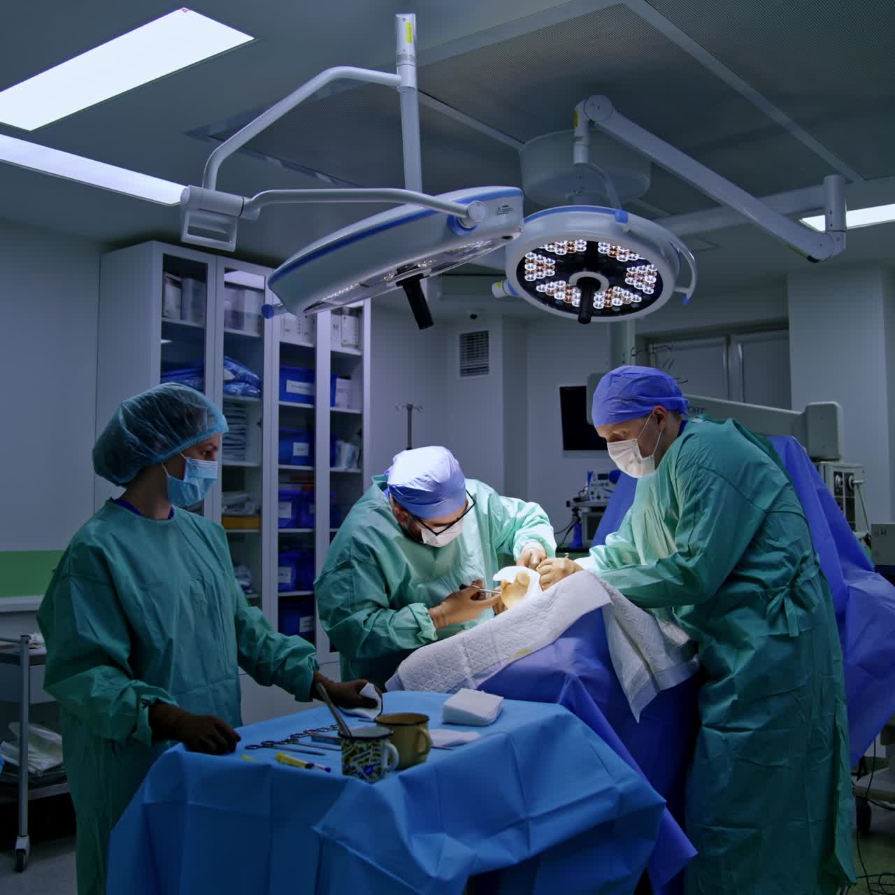 Main surgeon applies metal instrument at foot surgery. Assisting doctor holds the operated foot. Female nurse stands at table with tools