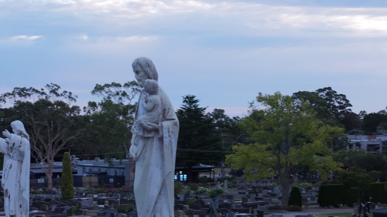 Patung yang tenang berdiri khidmat di pemakaman saat matahari terbenam di Ferntree Gully, Melbourne. Suasana yang damai diperkuat oleh nuansa keemasan lembut dari matahari terbenam.