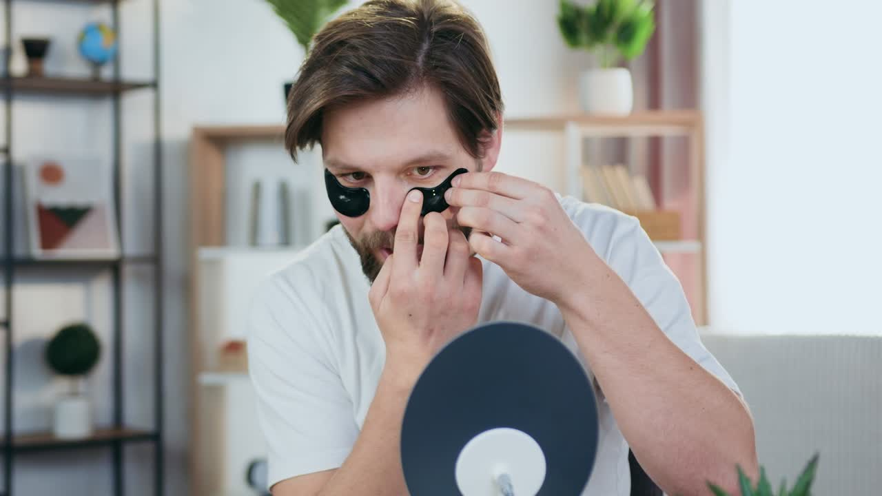 hombre joven y guapo sonriente y barbudo sentado frente a un pequeño espejo en casa y aplicando parches de colágeno negro en los ojos, concepto de piel facial