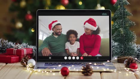 Happy-family-wearing-santa-hats-on-laptop-video-call,-with-christmas-decorations-and-tree