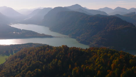 Aerial-view-pull-away-from-sunrise-glowing-over-hazy-Bavaria-mountain-forest-hills-and-Sylvenstein-lakeshore