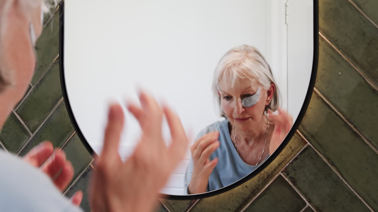 Senior woman applying eye patches while looking in bathroom mirror, at home