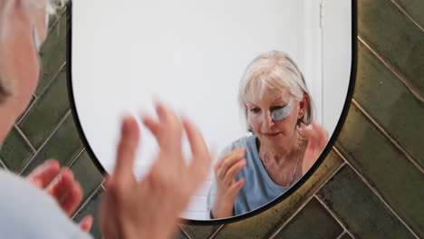 senior woman applying eye patches while looking in bathroom mirror, at home