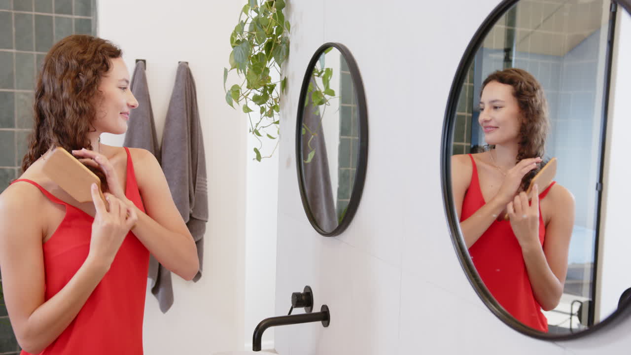 Brushing hair in bathroom, woman in red dress smiling at mirror reflection