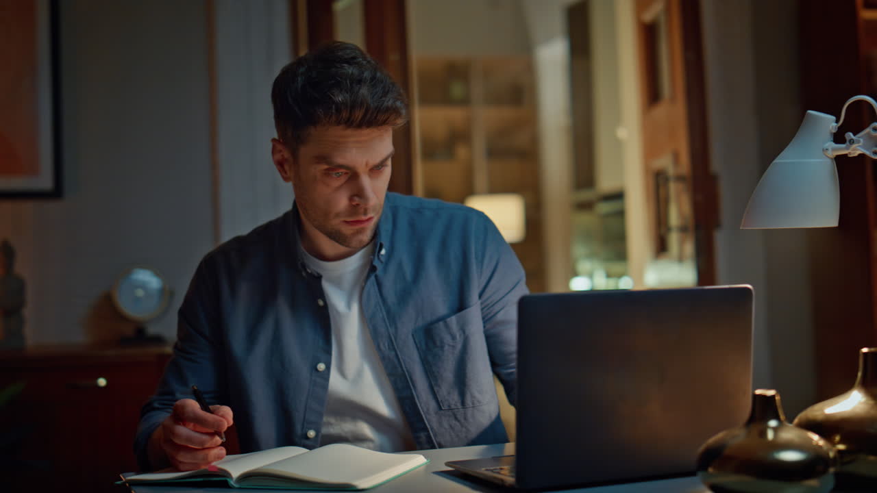Focused man working laptop at night apartment closeup. Serious guy writing notes