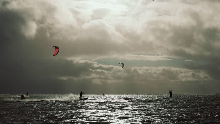 Slowmotion af kitesurfere, der rider på fladt, fyldigt vand under en storm, mens solen stråler gennem mørke skyer.