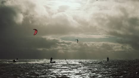 slowmotion of kitesurfers riding on flat, chubby water during a storm, while the sun beams through dark clouds