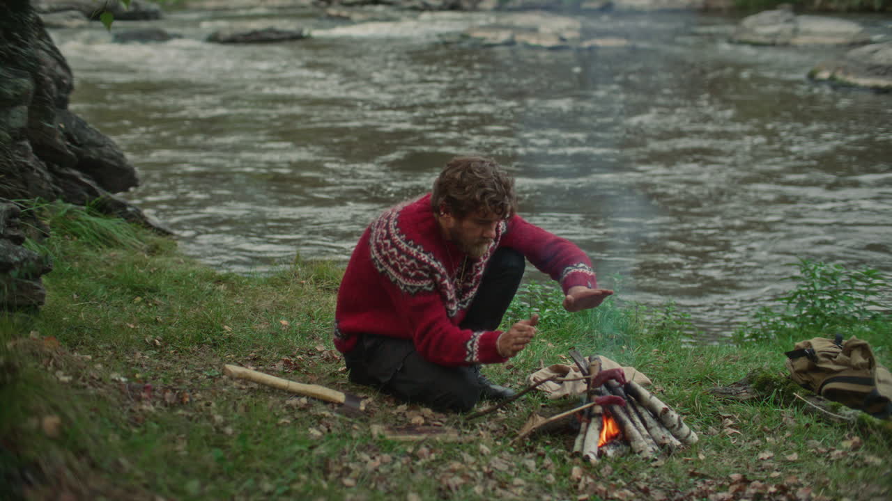 Survivalist Cooking Meat and Warming Hands over Campfire beside River