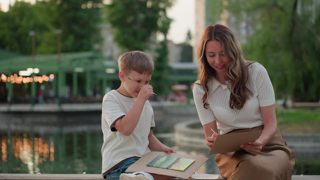 happy toddler holding paintbrush and board beside mom on wooden dock by pool under golden sunset glow, excitedly drawing and chatting while mother smiles and guides playful creation moment