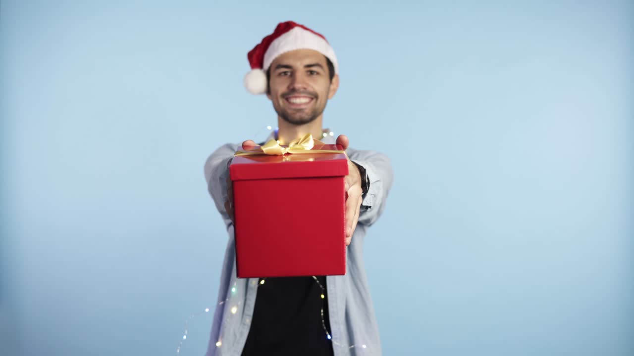 크리스마스 산타 모자와 화환을 목에 걸고 고립된 파란색 배경 위에 선물을 들고 손을 뻗은 채 행복한 얼굴을 하고 치아를 보여주는 자신감 넘치는 미소를 짓고 있는 청년