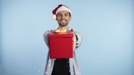 joven con gorro navideño de papá noel y guirnalda en el cuello sosteniendo un regalo sobre un fondo azul aislado con las manos extendidas y una cara feliz de pie y sonriendo con una sonrisa confiada mostrando los dientes