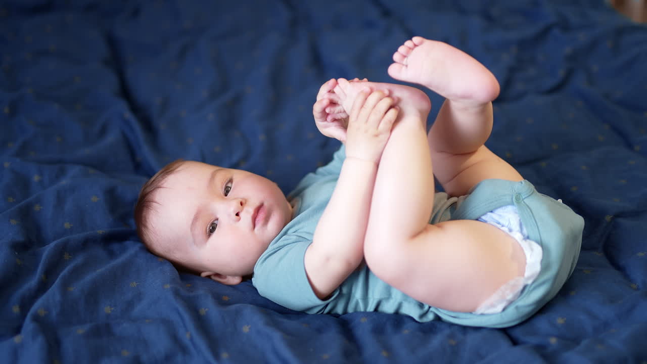 Sweet little Caucasian kid lies on back with his legs up. Beautiful baby boy plays with his bare feet. Blue cover backdrop.