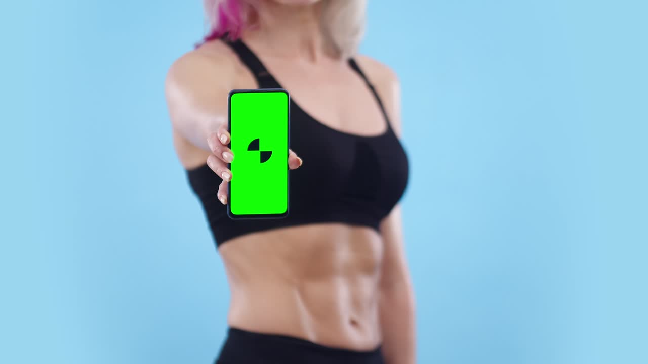 mujer con un vientre musculoso muestra el teléfono con una aplicación en el estudio, primer plano. mujer deportiva sostiene un teléfono en la mano, pantalla verde