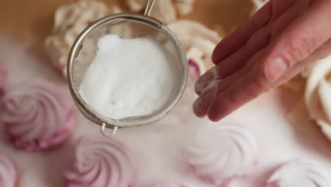 Close up of baker holding mesh sifter filled with fine sugar while gently sprinkling it over decorative cupcakes arranged on parchment, highlighting sweet detail and artistic dessert presentation