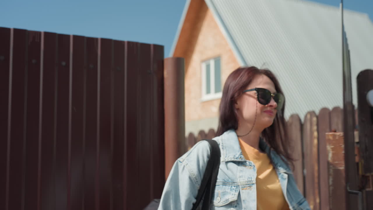 Young woman smiles warmly while closing brown wooden door after entering home on bright sunny day with house and fence in background