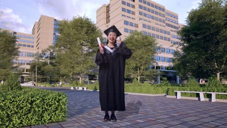 full body of asian woman student graduates in cap and gown holding diploma and making call me gesture in front of a magnificent university building