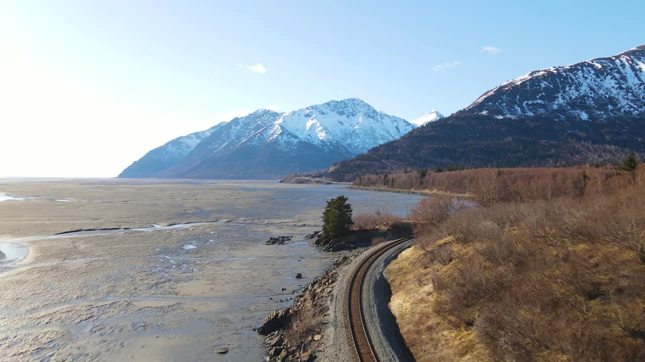화창한 날 배경에 눈 덮인 산이 있는 알래스카 비용을 따라 철로 위를 비행하는 4k 드론