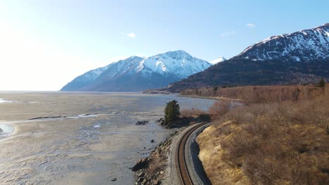 Dron-De-4k-Volando-Sobre-Las-Vías-Del-Tren-A-Lo-Largo-De-La-Costa-De-Alaska-Con-Montañas-Cubiertas-De-Nieve-En-El-Fondo-En-Un-Día-Soleado