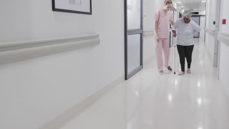 diverse female doctor helping senior female patient walking with stick in corridor, slow motion