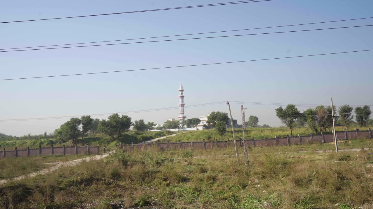 In a rural setting, there is a mosque with a tall tower, with a boundary wall enclosing the area.
